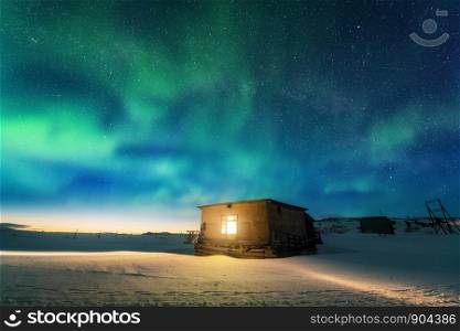 Aurora borealis over old small house with yellow light from the window. Northern lights in Teriberka, Russia. Blue sky with stars and green polar lights. Night winter landscape with aurora, building