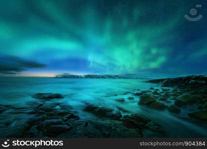 Aurora borealis over rocky beach and ocean. Northern lights in Teriberka, Russia. Starry sky with polar lights. Night winter landscape with aurora, sea with stones in blurred water, snowy mountains