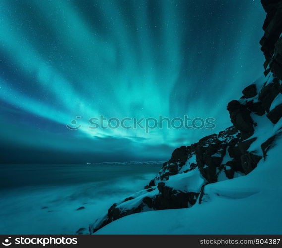 Aurora borealis over rocky beach and ocean. Northern lights in Teriberka, Russia. Starry sky with polar lights. Night winter landscape with aurora, sea with blurred water, snowy mountains. Travel