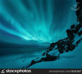 Aurora borealis over rocky beach and ocean. Northern lights in Teriberka, Russia. Starry sky with polar lights. Night winter landscape with aurora, sea with blurred water, snowy mountains. Travel