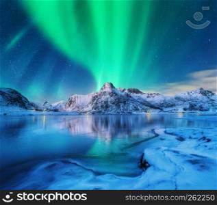 Aurora borealis over snowy mountains, frozen sea coast and reflection in water in Lofoten islands, Norway. Northern lights. Winter landscape with polar lights, ice in water. Starry sky with aurora. Aurora borealis over snowy mountains, frozen sea coast