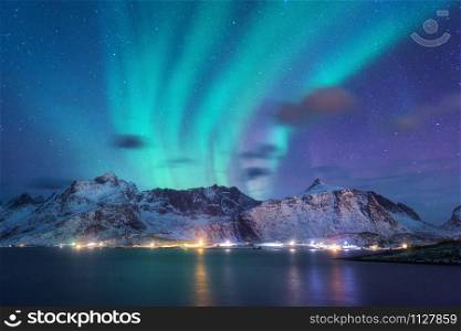 Aurora borealis over the sea, snowy mountains and city lights at night. Northern lights in Lofoten islands, Norway. Purple starry sky with polar lights. Winter landscape with aurora reflected in water