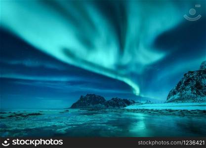 Aurora borealis over the snowy mountains and sandy beach in winter. Northern lights in Lofoten islands, Norway. Starry sky with polar lights. Night landscape with aurora, frozen sea coast, blue sky