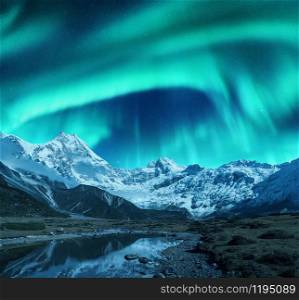 Aurora borealis over the snowy mountains, coast of the lake and reflection in water. Northern lights above snow covered rocks. Winter landscape with polar lights, lake. Starry sky with bright aurora