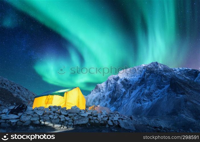 Aurora borealis, yellow glowing tent and snowy mountains. Northern lights, mountain range, starry sky at night in winter. Rocks, sky with stars and polar lights. landscape with green aurora. Space