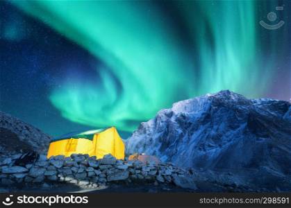 Aurora borealis, yellow glowing tent and snowy mountains. Northern lights, mountain range, starry sky at night in winter. Rocks, sky with stars and polar lights. landscape with green aurora. Space