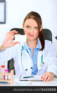 Authoritative medical doctor woman sitting at office table and holding blank business card&#xA;