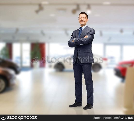 auto business, car sale and people concept - happy smiling businessman or dealer in suit over auto show background