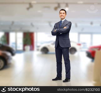 auto business, car sale and people concept - happy smiling businessman or dealer in suit over auto show background