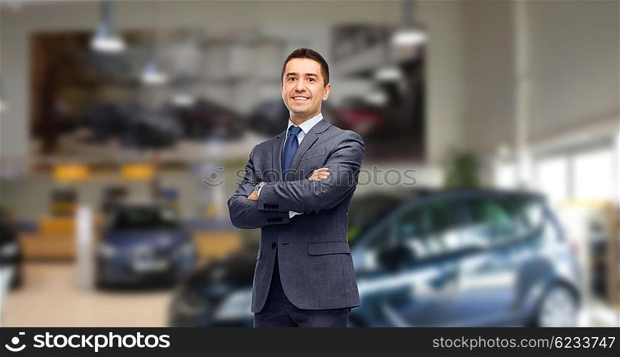 auto business, car sale and people concept - happy smiling businessman or dealer in suit over auto show background