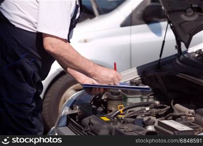 Auto mechanic checking car engine and writing on the clipboard. Car or motor mechanic checking a car engine and writing on the c