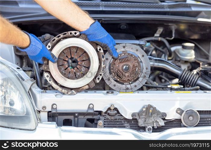 Auto mechanic holding used car pressure plate and clutch disc in front of the vehicle engine