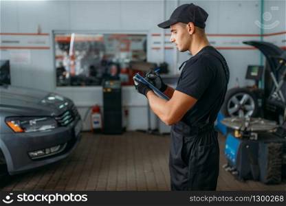 Auto mechanic in uniform holds inspection report, tire service. Worker repairs car tyre in garage, professional automobile inspection in workshop, vehicle on lift jack on background. Mechanic holds inspection report, tire service