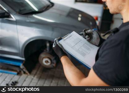 Auto mechanic in uniform holds inspection report, tire service. Worker repairs car tyre in garage, professional automobile inspection in workshop, vehicle on lift jack on background