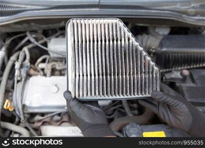 Auto mechanic wearing protective work gloves holding dirty used air filter above a car engine