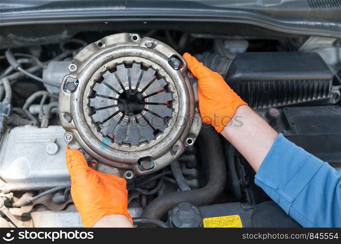 Auto mechanic wearing protective work gloves holding used clutch pressure plate above a car engine