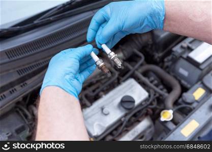 Auto mechanic wearing protective work gloves holds old and new spark plugs over a car engine