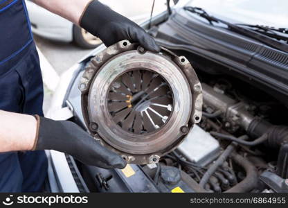 Auto mechanic wearing protective work gloves holds old clutch basket over a car engine