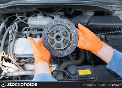 Auto mechanic wearing protective work gloves holds old clutch disc above a car engine