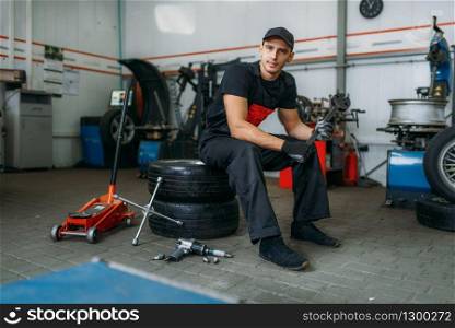 Auto mechanic with wrench sitting on wheels, tire service. Technician repairs car tyre in garage, professional automobile inspection in workshop. Auto mechanic sitting on wheels, tire service