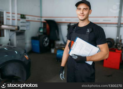 Auto repairman in uniform holds inspection report, tire service. Technician repairs car tyre in garage, professional automobile inspection in workshop, vehicle on lift jack on background