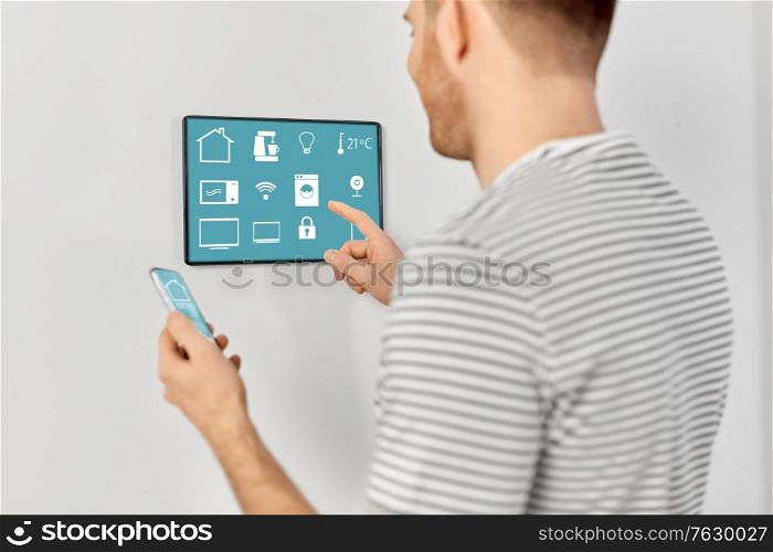 automation, internet of things and technology concept - close up of happy smiling man with smartphone looking at tablet pc computer on wall with menu icons on screen at smart home. man with smartphone and tablet pc at smart home