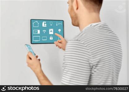automation, internet of things and technology concept - close up of happy smiling man with smartphone looking at tablet pc computer on wall with menu icons on screen at smart home. man with smartphone and tablet pc at smart home
