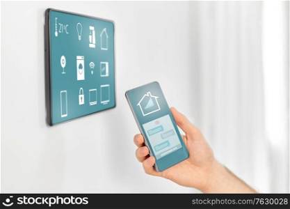 automation, internet of things and technology concept - close up of male hand with smartphone looking at tablet pc computer on wall with menu icons on screen at smart home. man with smartphone and tablet pc at smart home