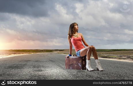 Autostop travel. Young pretty girl traveler sitting on suitcase aside of road