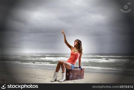 Autostop travel. Young pretty girl traveler sitting on suitcase aside of road