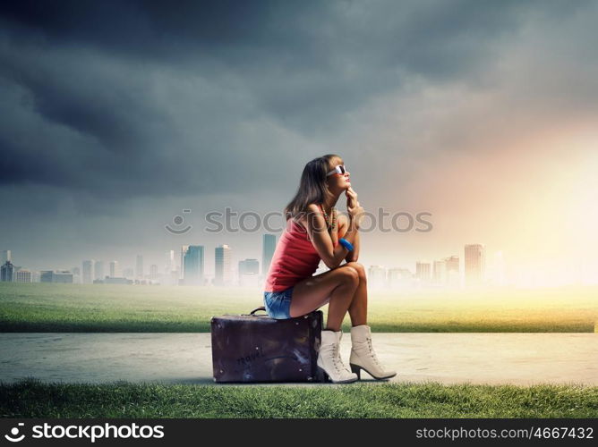 Autostop traveling. Young pretty girl traveler sitting on suitcase aside of road