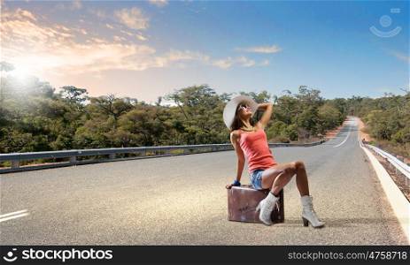 Autostop traveling. Young pretty girl traveler sitting on suitcase aside of road