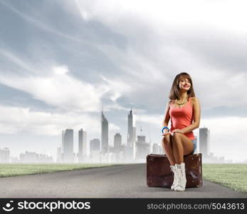 Autostop traveling. Young pretty girl traveler sitting on suitcase aside of road