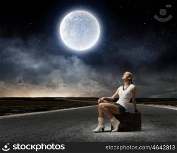 Autostop traveling. Young woman sitting on briefcase along roadside