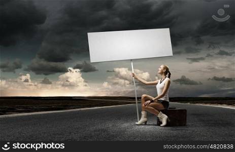Autostop traveling. Young woman with blank banner sitting on suitcase