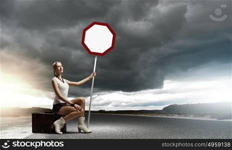 Autostop traveling. Young woman with road sign sitting on suitcase