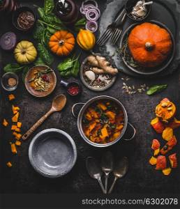 Autumn and winter cooking and eating with pumpkin dishes. Vegetarian stew in cooking pot with spoon and vegetables ingredients on dark kitchen table background, top view. Healthy seasonal food