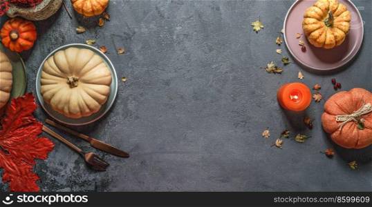 Autumn background frame with various pumpkins, red fall leaves, plates, cutlery and candle at dark grey backdrop. Seasonal fall table setting. Top view with copy space.