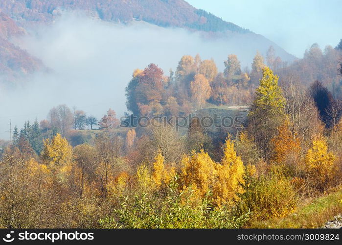 Autumn Carpathian Mountains landscape with multicolored trees and small hamlet on slope, and misty cloud over (Rakhiv district, Transcarpathia, Ukraine).