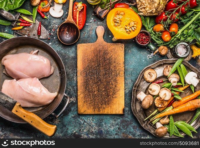 Autumn cooking with organic harvest vegetables, pumpkin and chicken on rustic kitchen table background with empty cutting board, top view, frame