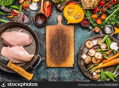 Autumn cooking with organic harvest vegetables, pumpkin and chicken on rustic kitchen table background with empty cutting board, top view, frame