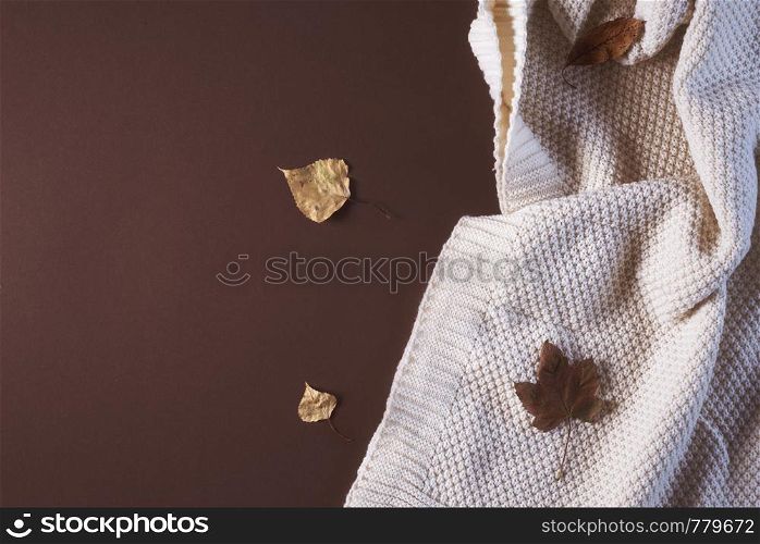 Autumn coziness concept with a white knitted wool blanket and colorful fallen leaves on a brown paper background. Above view of cozy autumn settings.