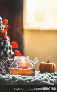 Autumn cozy still life with homemade tea in glass pot with steam on wool blanket at blurred background. Seasonal hot beverage in fall at home. Front view.