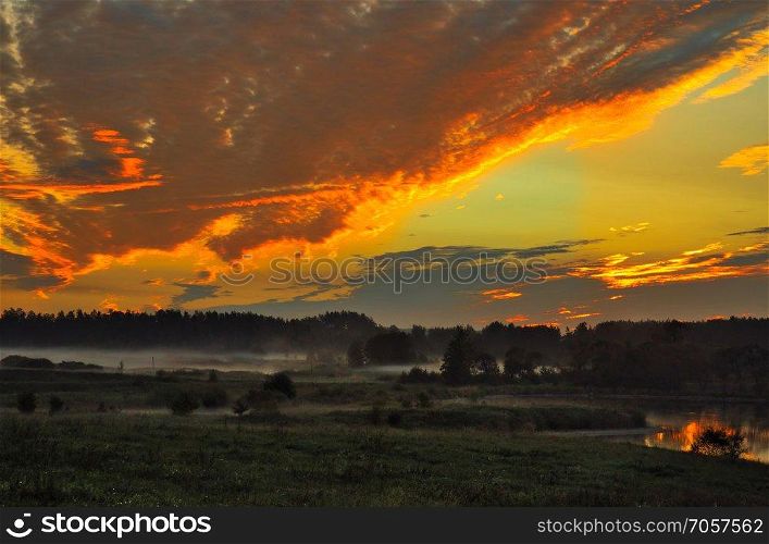 Autumn dawn over the meadow and pond. Beautifully highlighted in red and yellow clouds and light fog over the meadow. Mysterious, moody landscape. Mazury district in Poland.Horizontal view