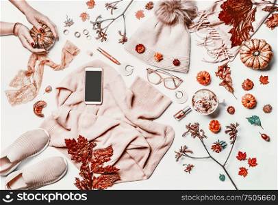 Autumn fashion woman clothes and shoes flat lay layout with smartphone mock up screen, sweater, knitted hat, scarf, eyeglasses, cosmetics,pumpkin, cappuccino and fall leaves. Female hands.White desk