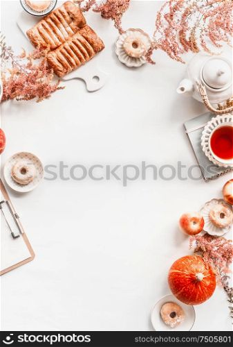 Autumn flat lay layout with copy space: tea setting, cakes, pastry and donuts with autumn flowers arrangement and pumpkin on white desktop background. . Top view. Frame