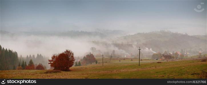 Autumn fog in mountain village. Cloudy rainy misty autumn day panorama. Mountain hills in clouds of fog