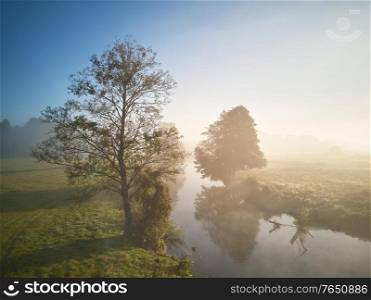 Autumn foggy sunrise. Colorful misty morning dawn. Fall rural scene. Small river with trees in meadow and field, Belarus
