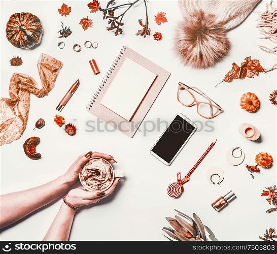Autumn home office. Woman hands with cup of cappuccino on white desktop with autumn objects: knitted hat,pumpkin, autumn leaves and flowers, smartphone mock up screen, eyeglasses and notebook
