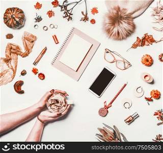 Autumn home office. Woman hands with cup of cappuccino on white desktop with autumn objects: knitted hat,pumpkin, autumn leaves and flowers, smartphone mock up screen, eyeglasses and notebook